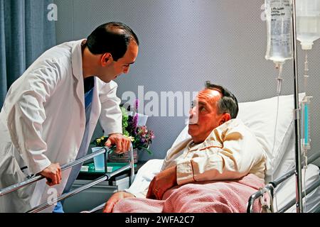 Un uomo in un letto d'ospedale impegnato in una conversazione con un medico che indossa un cappotto bianco. Foto Stock