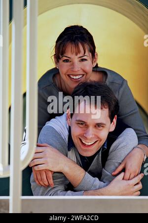 Un uomo e una donna gioiosi si abbracciano mentre sbirciano attraverso un tunnel del parco giochi, indossando abiti casual e condividendo un momento di felicità Foto Stock
