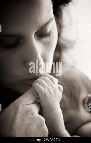 Una fotografia in bianco e nero ero a una giovane madre attraente che baciava delicatamente la mano del suo neonato su uno sfondo bianco Foto Stock