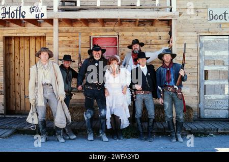 Un gruppo di sette persone si trova in fila, posando in costumi specifici del periodo rappresentativo dell'era del selvaggio West americano, di fronte a un saloon rustico Foto Stock