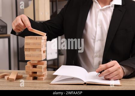 Giocare a Jenga. Edificio per uomini d'affari con blocchi di legno e scrittura del risultato in un notebook al tavolo all'interno, primo piano Foto Stock