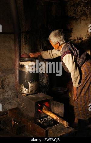 Una donna bhutanese anziana all'interno della sua cucina rustica versa acqua in un grande contenitore sopra il fuoco per preparare il tè caldo al burro di yak a Ogyen Choling vil Foto Stock