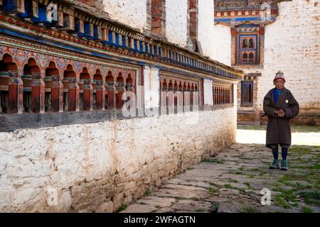 Un vecchio animato si trova di fronte alle ruote di preghiera in un antico tempio buddista; il villaggio di Ogyen Choling, la valle di Bumthang, il Bhutan, l'Asia Foto Stock