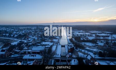 Questa immagine aerea presenta una tranquilla mattinata invernale su un'area suburbana, con un canale che attraversa il cuore del quartiere. L'alba dona un caldo bagliore alle fresche tonalità blu delle case e delle strade innevate. Il canale, parzialmente congelato, riflette la luce del sole del mattino presto, migliorando la tranquillità della scena. Alba invernale sul Canale Suburban. Foto di alta qualità Foto Stock