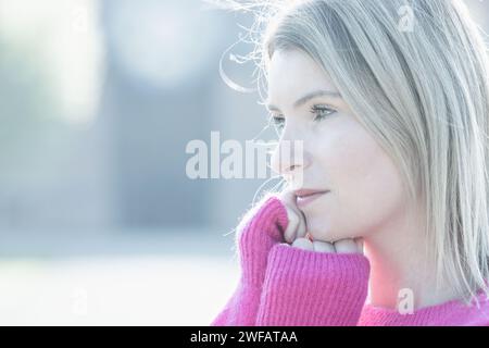Il ritratto ravvicinato raffigura una giovane donna caucasica in un morbido maglione rosa pastello. L'immagine è immersa in una luce fredda e diffusa, evidenziando l'umore pensivo sul suo viso. Il suo sguardo è diretto fuori dalla fotocamera, suggerendo un pensiero profondo o una riflessione. L'illuminazione ad alta luminosità accentua la sua carnagione discreta e la texture raffinata del maglione, mentre lo sfondo rimane indistinto, enfatizzando il suo momento introspettivo. Atmosfera pensiva in rosa pastello. Foto di alta qualità Foto Stock