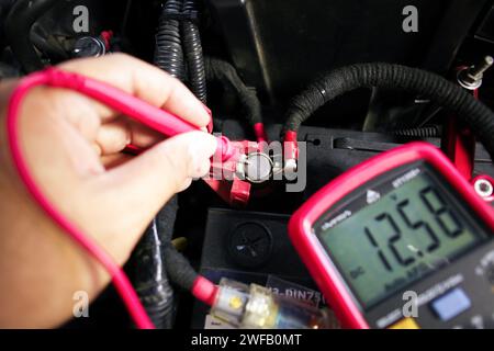 Test della tensione della batteria del veicolo con un multimetro digitale da parte di un tecnico automatico, visualizzazione della corrente di tensione del veicolo su schermo nero del multimetro digitale, manutenzione del veicolo Foto Stock