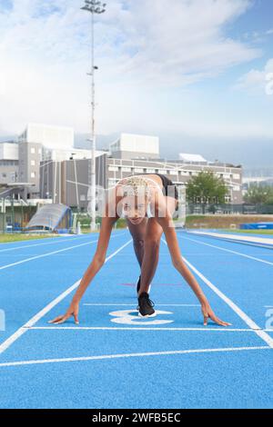 Un'atleta afroamericana si trova sulla linea di partenza della pista blu olimpica, incarnando il concetto di allenamento e dedizione da gara Foto Stock