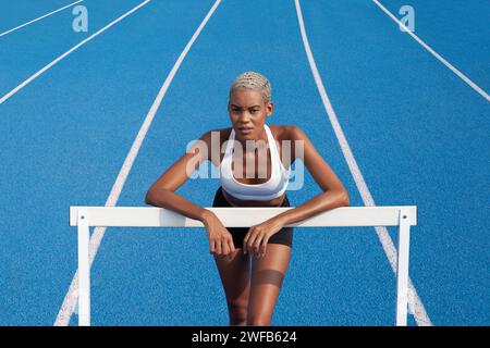 Un'atleta afroamericana si sta allenando sulla pista olimpica, appoggiandosi contro gli ostacoli, con corsie blu sullo sfondo, concetto di gara t Foto Stock