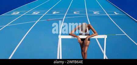 Un'atleta afroamericana si sta allenando sulla pista olimpica, appoggiandosi contro gli ostacoli, con corsie blu sullo sfondo, concetto di gara t Foto Stock