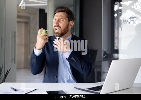 Un giovane uomo d'affari soffre di un forte mal di gola, siede a una scrivania in ufficio e gli ruba un aerosol medico in bocca. Foto Stock