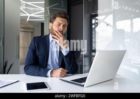 Un giovane uomo d'affari stanco seduto in ufficio alla scrivania, sbadiglia, si copre la bocca con la mano, si sente sonnolento. Foto Stock