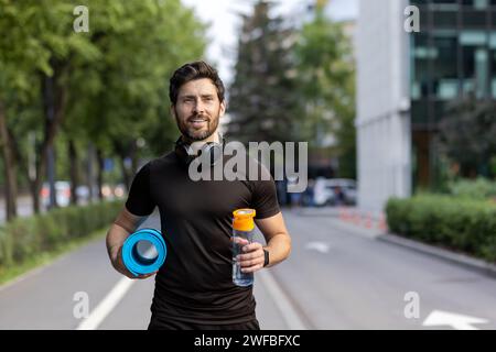 Uomo attivo con cuffie e tappetino yoga che cammina in ambiente urbano, tenendo una bottiglia d'acqua, trasuda salute e benessere. Foto Stock