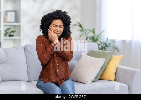 La giovane donna afroamericana sconvolta è a casa, seduta sul divano e chiusa gli occhi, tenendo la guancia con le mani, avendo un forte mal di denti, avendo bisogno dell'aiuto di un dentista. Foto Stock