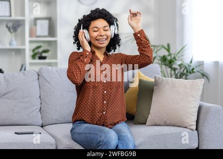 Giovane donna gioiosa che si diverte a ascoltare musica, ballare sul divano a casa, sentire il ritmo ad occhi chiusi, intimo interno del salotto. Foto Stock