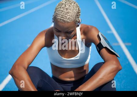 Atleta donna dopo aver eseguito gli esercizi sportivi, indossa auricolari e smartphone con fascia da braccio. Donna afroamericana seduta su pista olimpica con corsia blu Foto Stock