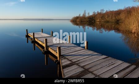 Molo di legno ghiacciato all'alba di fronte a un lago liscio a specchio. Vista meditativa sull'acqua calma fino all'orizzonte. Foto Stock