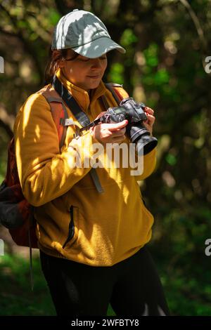 Bella donna latinoamericana che cammina con uno zaino rosso e un berretto blu che rivede le foto sulla sua fotocamera nel mezzo di una foresta durante una soleggiata festa Foto Stock