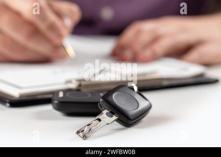 Chiavi dell'auto sulla scrivania con l'uomo che firma i documenti di acquisto sullo sfondo. Primo piano delle moderne chiavi nere dell'auto completando a mano la polizza assicurativa o renta Foto Stock