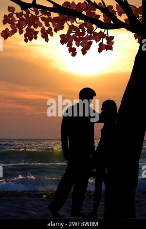 Gli amanti della coppia si stagliano sotto un albero appoggiato al tronco dell'albero in riva al mare al tramonto Foto Stock