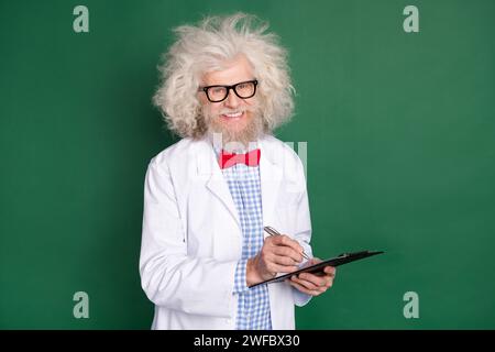 Foto di un simpatico e allegro scienziato dell'età che indossa occhiali a farfalla con cappotto bianco che scrive carte sorridenti di colore verde isolato Foto Stock