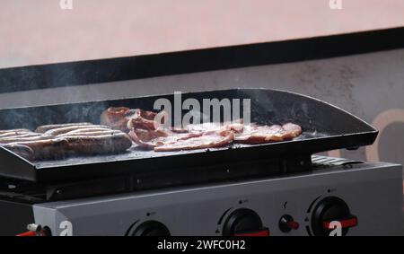 Pancetta di cottura e salsicce su un piano cottura a gas portatile. Foto Stock