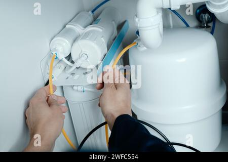 primo piano di un uomo caucasico, che indossa tute blu, che installa un sistema di osmosi inversa sotto il lavandino di una cucina Foto Stock