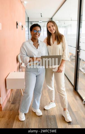 Due diverse colleghi donne utilizzano un computer portatile, in piedi in ufficio. Hanno in mano un laptop, guardano la fotocamera e sorridono, lunghezze intere, atmosfera da ufficio cordiale e concetto di lavoro di squadra Foto Stock