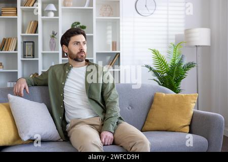 Uomo seduto sul divano a casa, serio e con una camicia casual, ragazzo maturo in salotto che si rilassa di giorno. Foto Stock