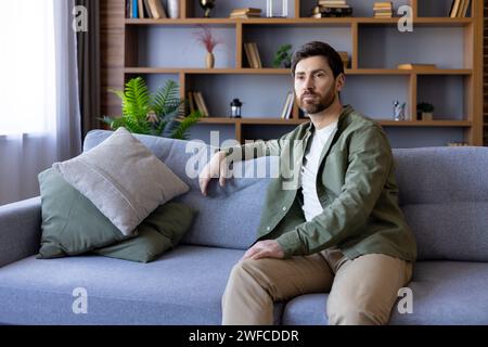 Uomo seduto sul divano a casa, serio e con una camicia casual, ragazzo maturo in salotto che si rilassa di giorno. Foto Stock