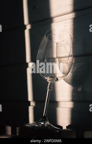 vetro rotto alla luce del sole su fondo di legno. Bicchiere da vino danneggiato con bordi affilati. Vetro incrinato nelle ombre. Natura morta creativa. Oggetto fragile Foto Stock