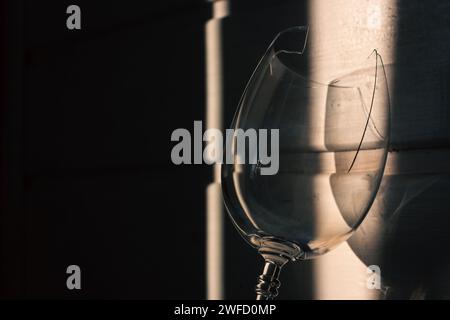 vetro rotto alla luce del sole su fondo di legno. Bicchiere da vino danneggiato con bordi affilati. Vetro incrinato nelle ombre. Natura morta creativa. Oggetto fragile Foto Stock