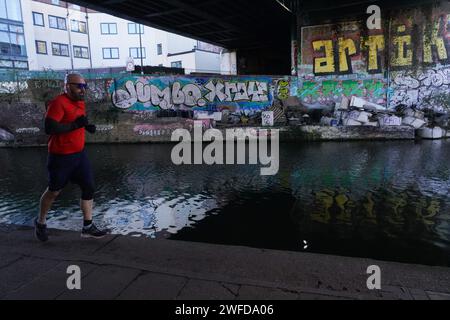 Un uomo corre lungo il sentiero di traino vicino al Regent's Canal sotto un ponte al sole di prima mattina d'inverno. Le pareti sono coperte di grafitti. Foto Stock