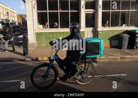 Un uomo Deliveroo cavalca la sua bici al sole invernale di prima mattina vicino al mercato di Broadway a Hackney, Londra, Regno Unito. Foto Stock