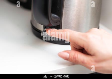 Il dito di una donna che preme l'interruttore di accensione sul bollitore elettrico. Messa a fuoco selettiva. Lo spazio di copia è sul lato destro. Foto Stock