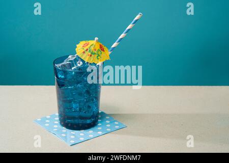 Cocktail in bicchiere blu riempito di ghiaccio con piccolo ombrello giallo e cannuccia su tovagliolo blu su un tavolo bianco su sfondo blu Foto Stock