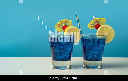 Due cocktail blu in un bicchiere pieno di ghiaccio guarnito con fette di limone, ciliegie, ombrelli gialli e sorbetti su un tavolo bianco contro una schiena blu Foto Stock