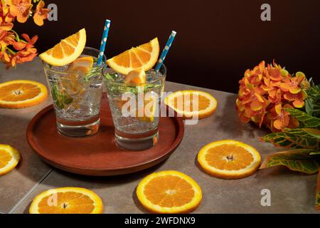 Due cocktail in un bicchiere pieno di ghiaccio e foglie di menta guarnite con una fetta d'arancia e sorbetto su un piatto di legno su un tavolo di ceramica con ora Foto Stock