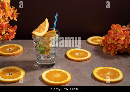 Cocktail in vetro pieno di ghiaccio e foglie di menta guarnite con fette d'arancia e sorbetto su un tavolo di ceramica con fette d'arancia contro il rosso scuro Foto Stock