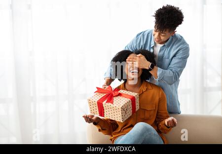 Un giovane afro-americano sorridente ha chiuso gli occhi alla signora, regala una confezione regalo all'interno del soggiorno Foto Stock