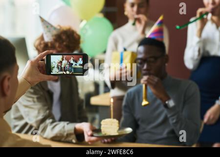 Mano di un giovane che tiene lo smartphone davanti a un gruppo di colleghi felici che festeggiano il compleanno di un lavoratore d'ufficio Foto Stock