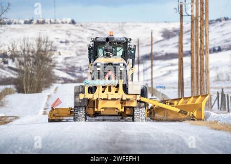 Rocky View County Alberta Canada, 24 gennaio 2024: Una livellatrice ara la neve su una strada di ghiaia rurale mentre passa pali telefonici in legno lungo il Canada Foto Stock