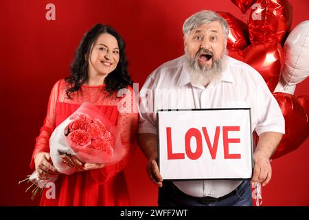 Bella coppia matura scioccata con palloncini a forma di cuore, bouquet di rose e AMORE per le parole su sfondo rosso. Festa di San Valentino Foto Stock