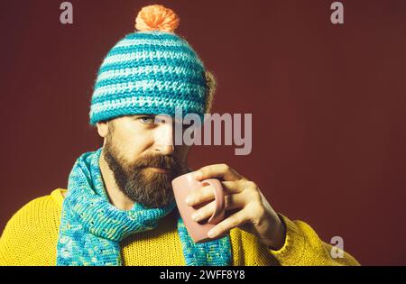 Bell'uomo barbuto in abiti alla moda che beve bevande dalla tazza. Uomo serio ed elegante in maglione e cappello avvolto in sciarpa con tazza di caldo Foto Stock