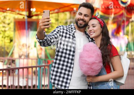 Felice giovane uomo e la sua ragazza con caramelle di cotone che fanno selfie a Funfair Foto Stock