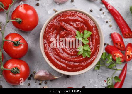 Composizione piatta con ketchup organico in recipiente su un tavolo grigio testurizzato. Salsa di pomodoro Foto Stock