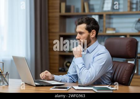 Uomo barbuto concentrato che guarda con attenzione lo schermo del computer portatile e tiene la mano sotto il mento nello spazio di lavoro. Il proprietario di una piccola impresa controlla gli estratti conto mensili online e le fatture contabili. Foto Stock