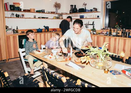 Madre sorridente durante la cena con figli seduti al tavolo da pranzo in cucina Foto Stock