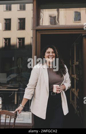 Donna d'affari felice che tiene una tazza di caffè in piedi vicino all'ingresso dell'ufficio Foto Stock