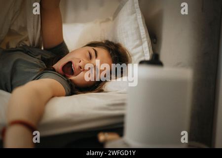 Ragazza che sbadiglia sul letto mentre si sveglia la mattina a casa Foto Stock