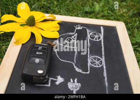 lavagna nera con un'auto elettrica disegnata presso una stazione di ricarica, chiave dell'auto e un fiore giallo, immagine simbolo Foto Stock